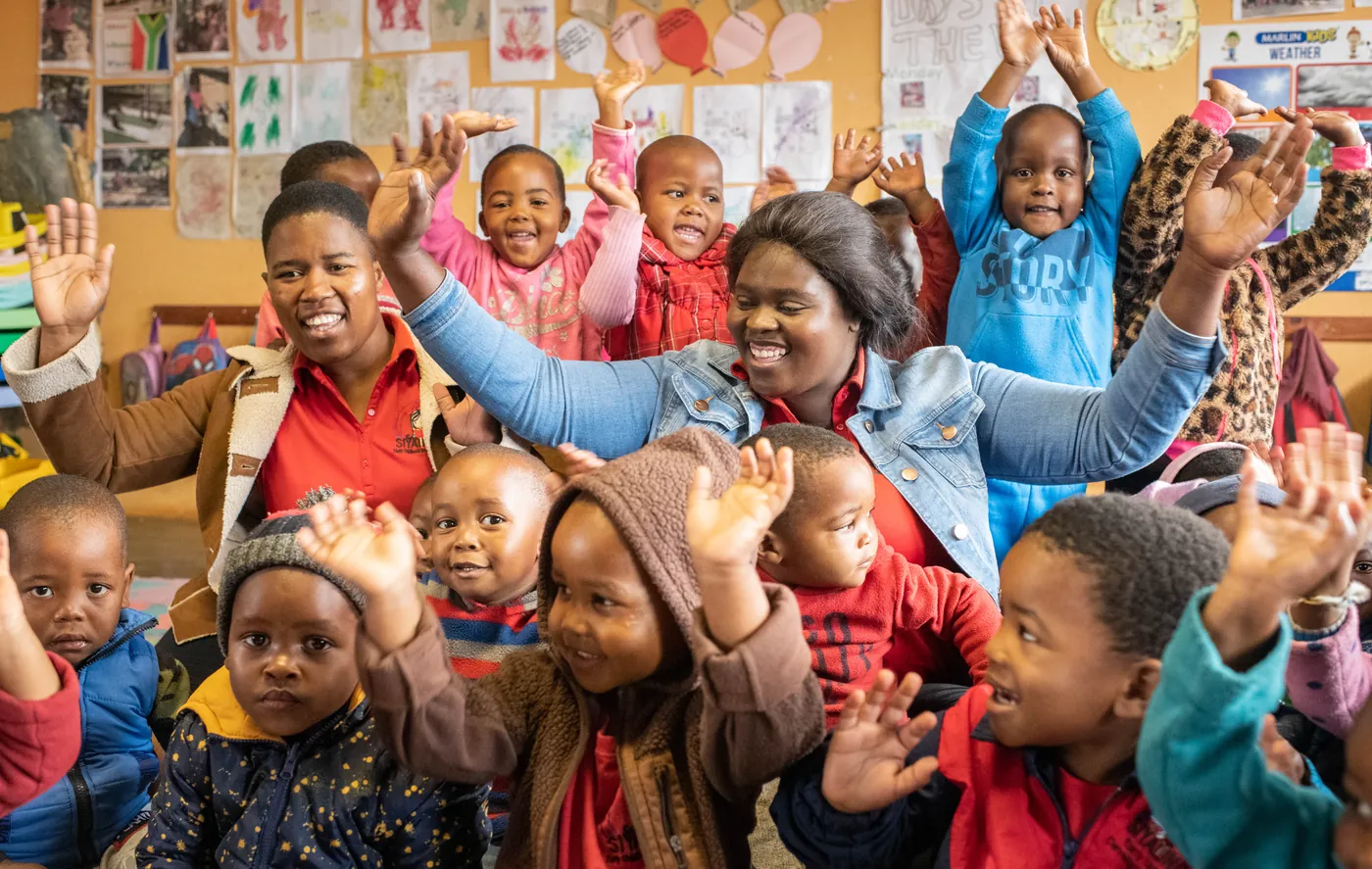 A Siyabonga teacher and children with their arms raised in the classroom