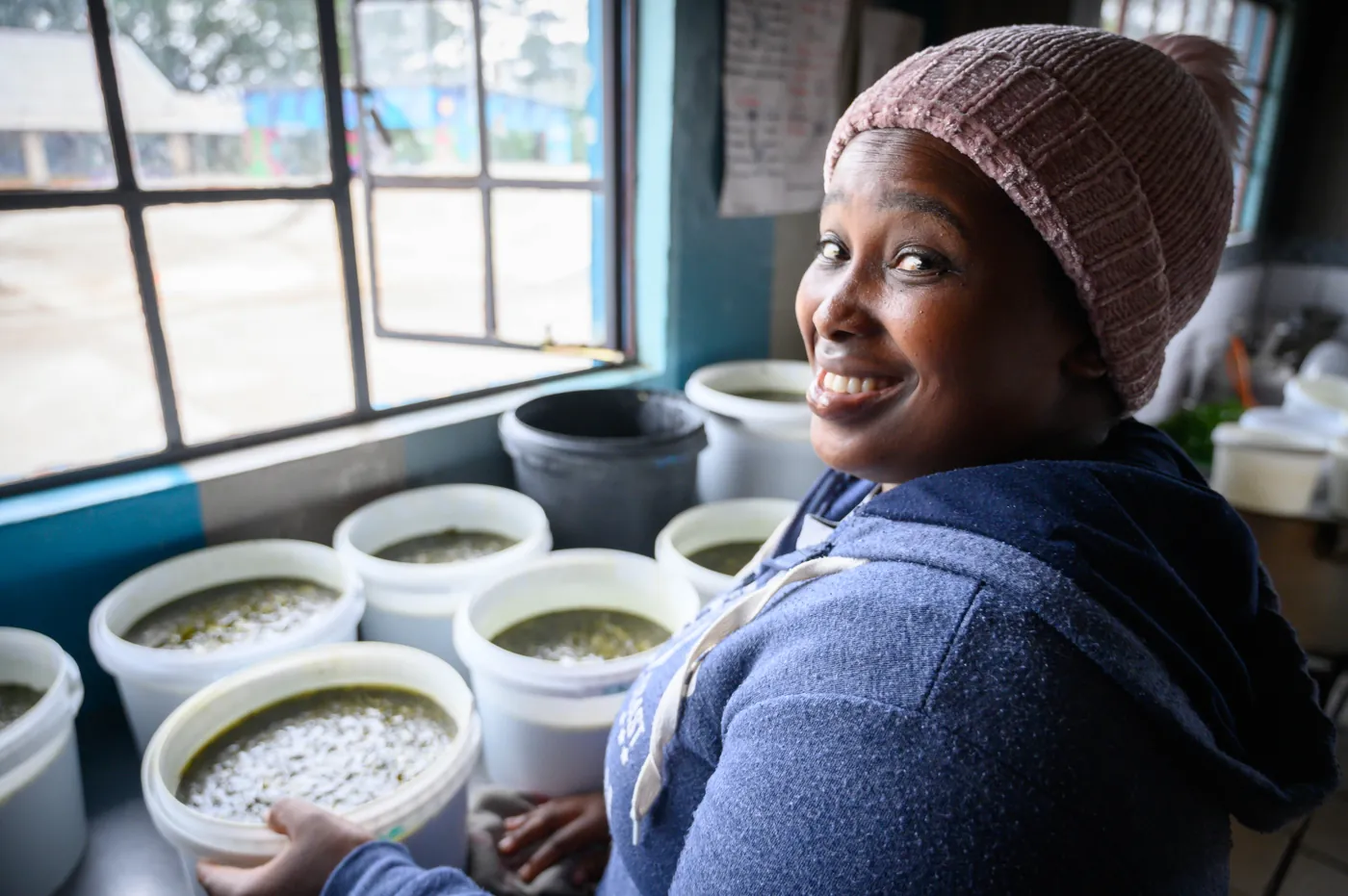 A Thanda cook smiling beside buckets of porridge in the kitchen