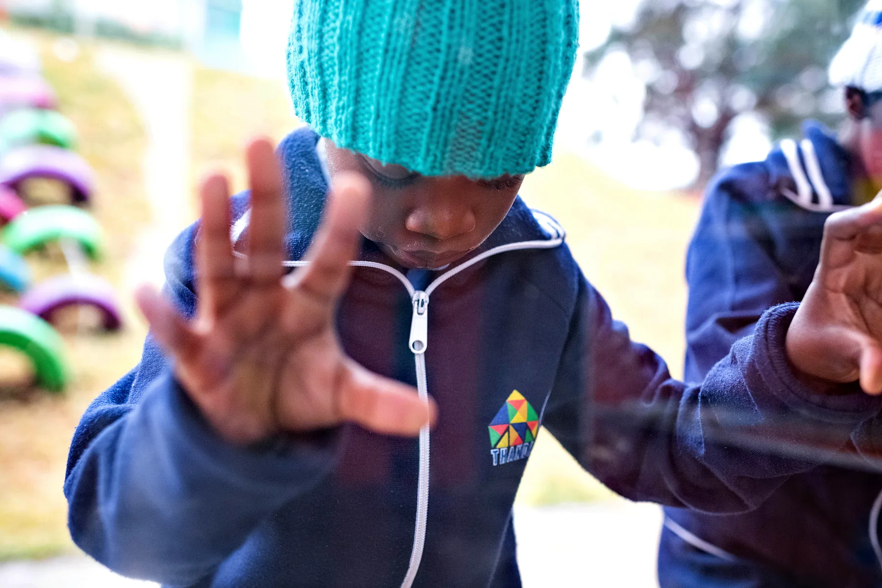 A boy in a Thanda hoodie and teal beanie pressing his hand to the window