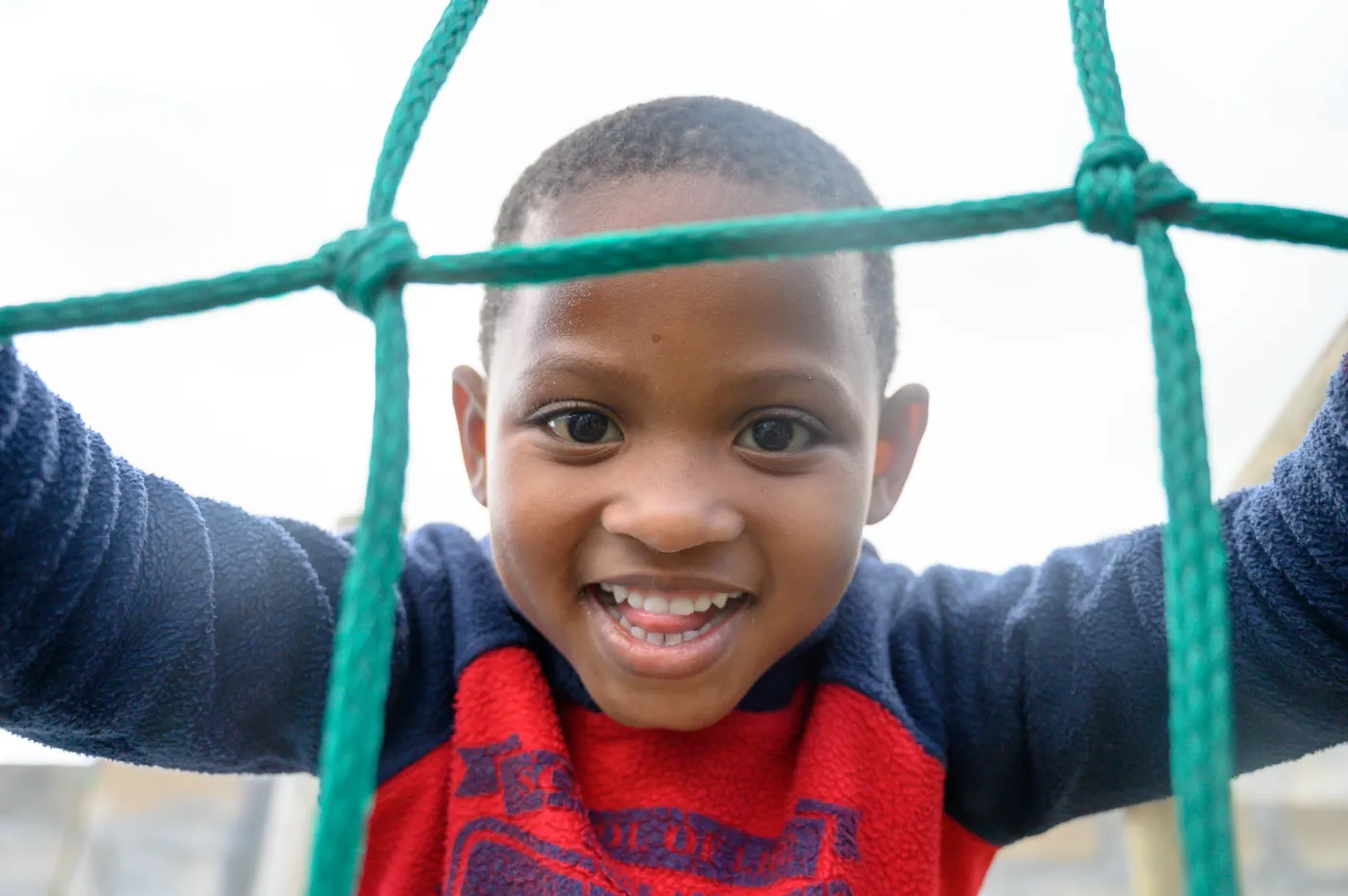A boy smiling through a climbing net on a True North playground